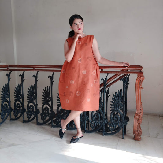 Wearing an orange sleeveless dress with hand-embroidered abstract motifs.